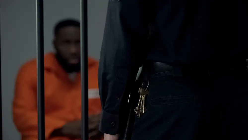 Agent pénitentiaire en uniforme de surveillant portant un trousseau de clés face à un détenu derrière des barreaux.