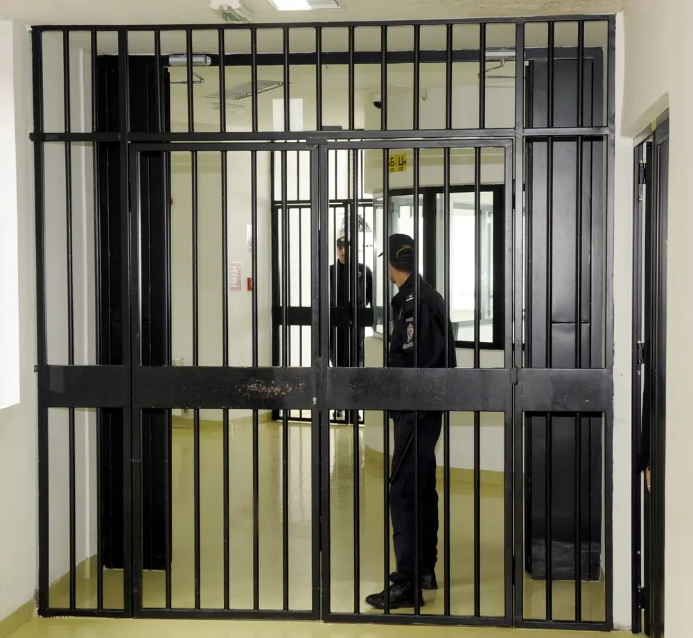 Agent pénitentiaire en uniforme de surveillant derrière une grille de sécurité dans une prison belge.