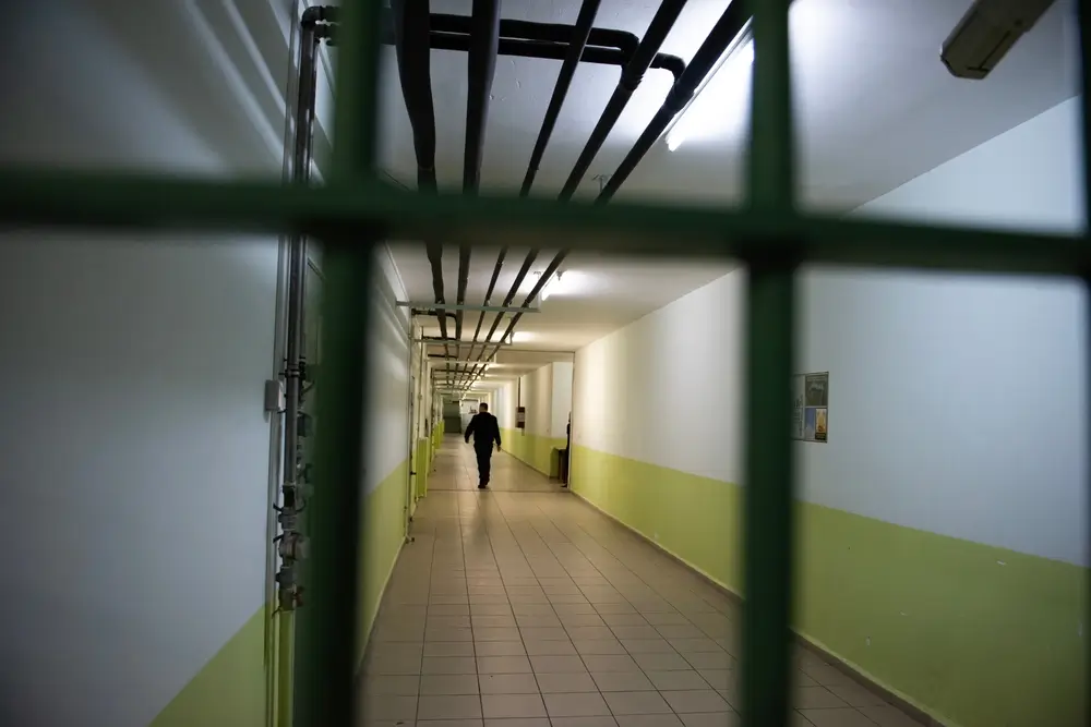 Agent pénitentiaire effectuant une ronde de surveillance dans un couloir de prison, aperçu à travers des barreaux.