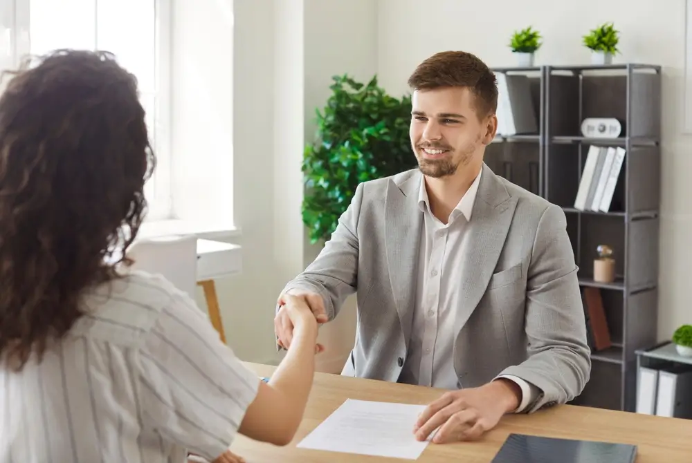 Candidat souriant en costume gris serrant la main d'une recruteuse lors d'un entretien de recrutement.