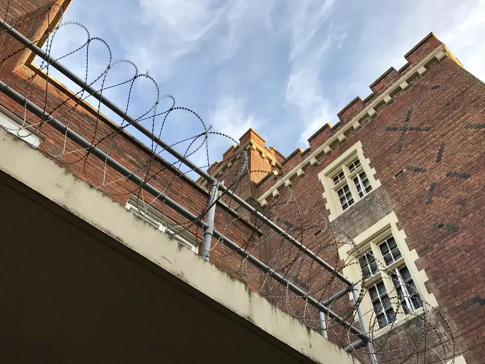 Mur d'enceinte d'un établissement pénitentiaire avec fils barbelés et bâtiment en briques sous un ciel bleu.